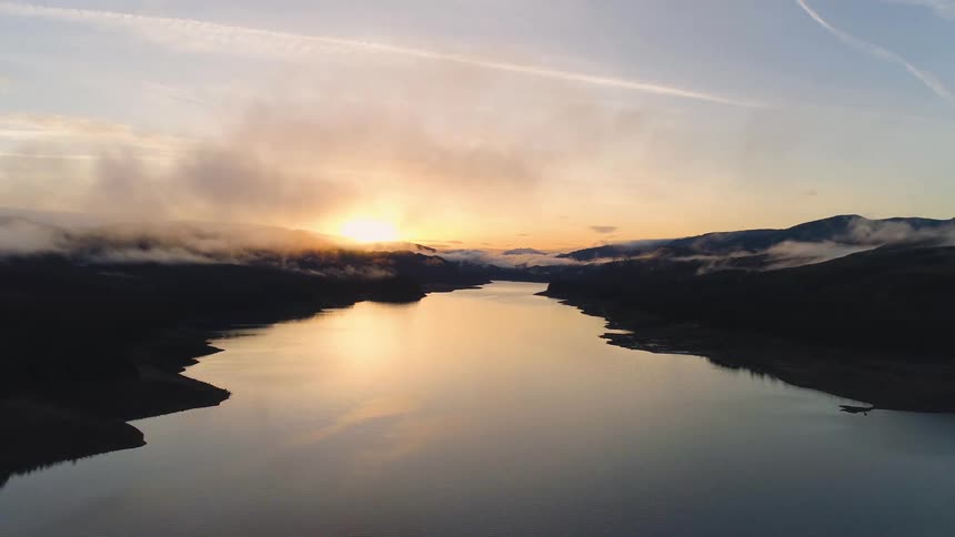 A stunning high aerial shot glides forward over a serene river that perfectly reflects the golden sunset winding its way through dark silhouetted mountains shrouded in lowhanging clouds on the horizon