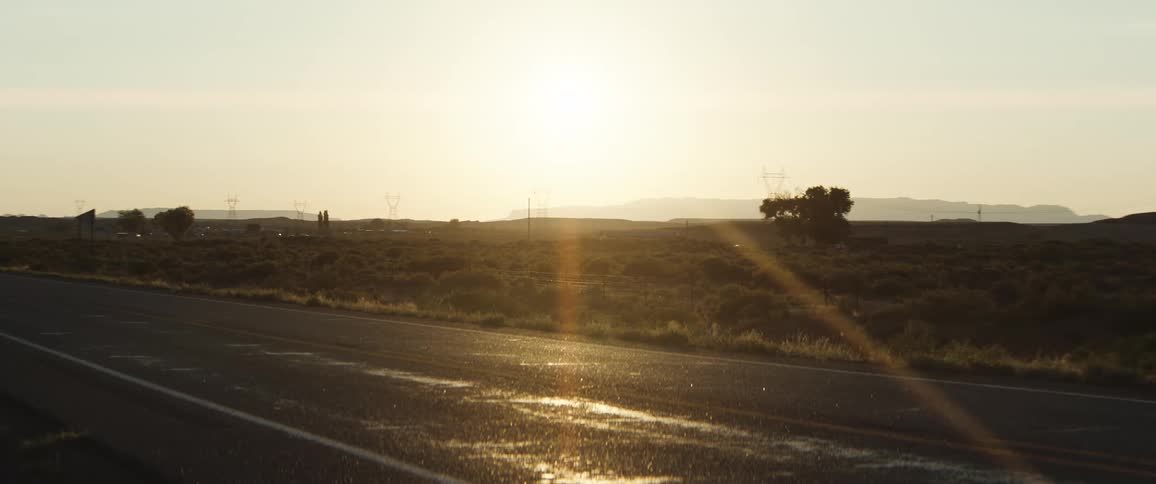 The sun shines brightly over a desert highway casting a lens flare and illuminating wet patches on the asphalt