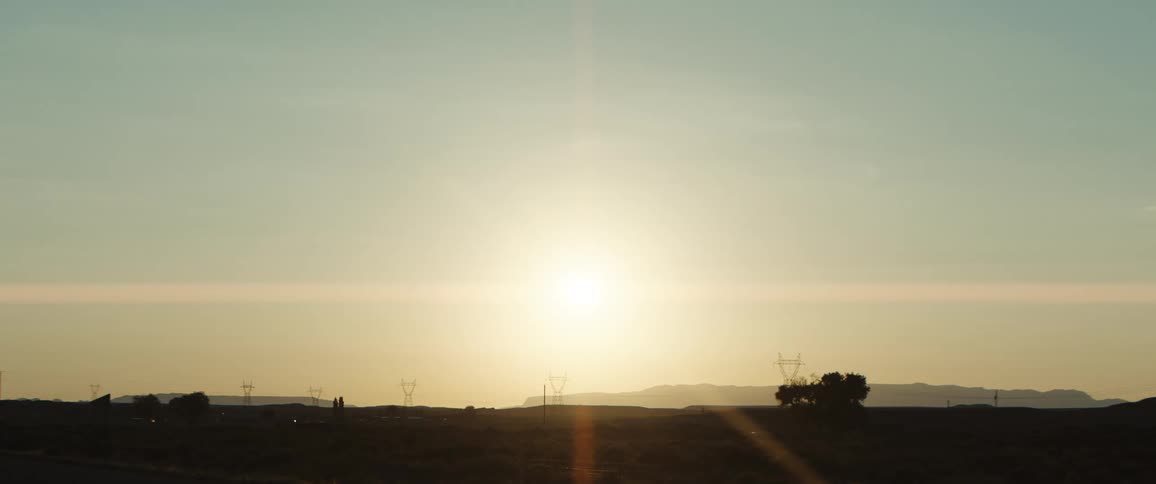 A golden sun sets over a vast arid landscape with distant mountains power lines and a few scattered trees