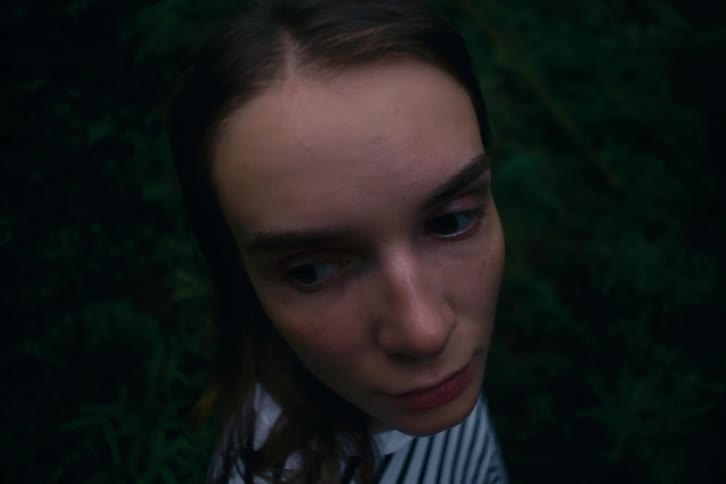 A woman peers upward then downward with tense wary eyes and a tight mouth her expression small and alert as if startled by something offframe