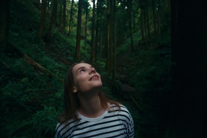 A young woman with an expressive face looks up at the towering trees and lush undergrowth of a dense forest