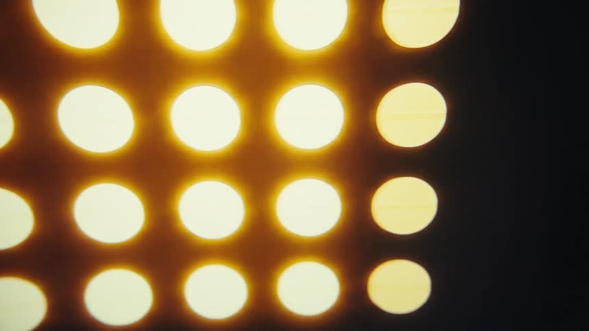 A closeup shot shows a wall of circular stage lights that are blinking on and off in a pattern the lights are a warm golden color and are arranged in a gridlike formation