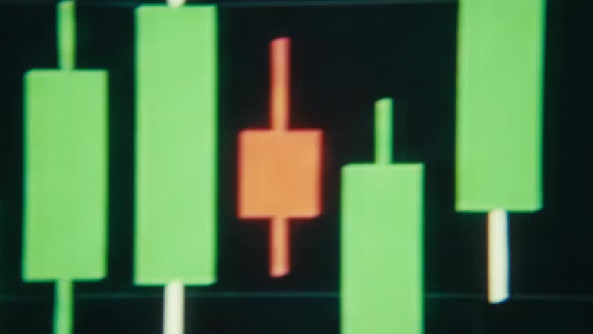 A monitor displays a stock market candlestick chart with green and red candles moving across a dark grid