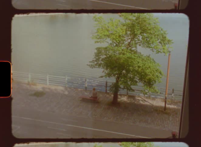 The scene is captured through an oldfashioned film reel effect with visible borders showing a static shot of a person riding a bicycle down a path next to a body of water a large green tree stands in the center of the frame with sunlight casting shado