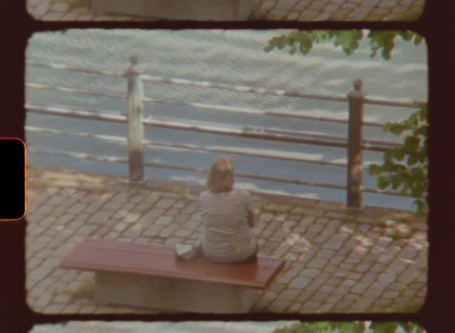 The woman eats while sitting on a bench by the water