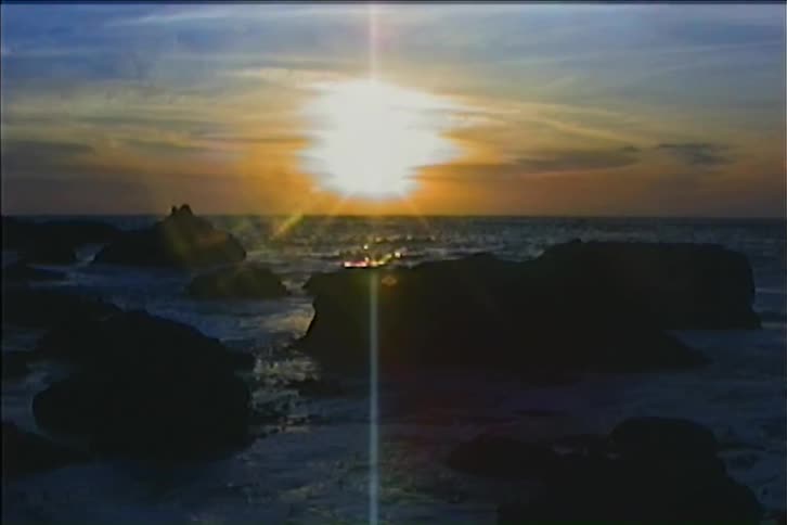 The sun sets over a rocky coastline casting a bright lens flare and illuminating the ocean waves
