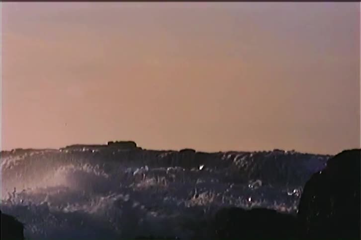 This vintage video captures a large wave crashing against a dark rocky shore the foamy white water explodes upward against a soft muted sky and the camera quickly pans down to follow the waves retreat