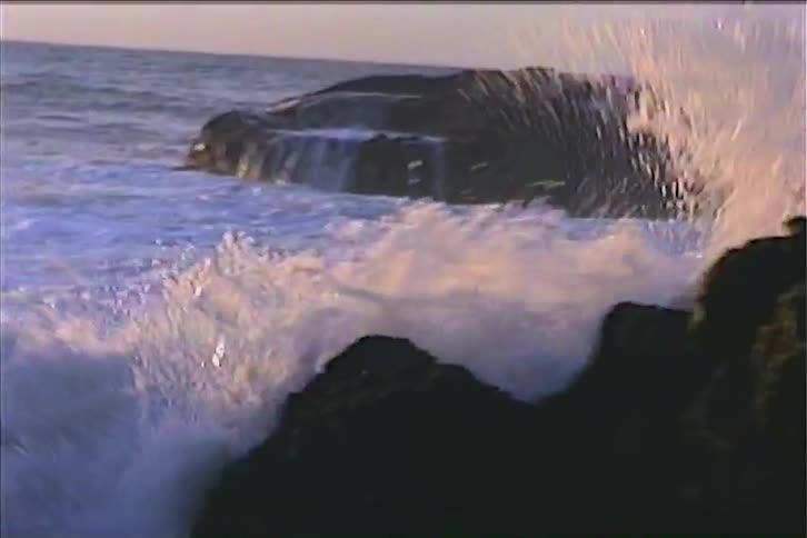 The video begins with a wide shot of large ocean waves crashing over a rocky coastline the camera then zooms in on the waves as they violently break against the dark jagged rocks creating a spray of white foam and water