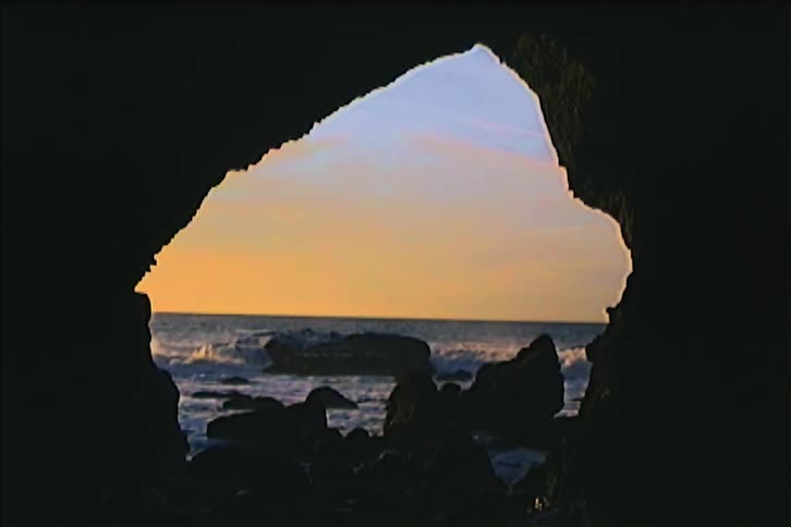 This static shot shows the ocean and a rocky beach from inside a cavelike opening the waves crash against the rocks in the distance framed by the dark silhouette of the rock formation