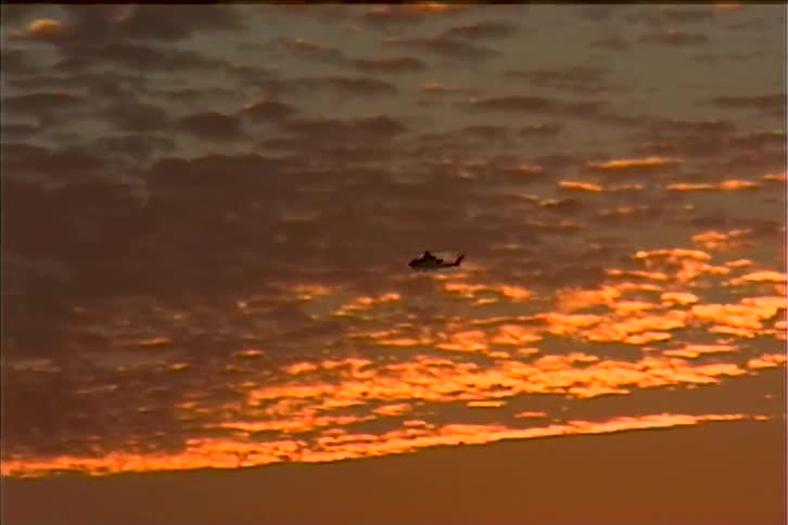 The first line of the video shows a vibrant sunrise over the ocean with the suns reflection visible as a small glowing dome on the waters surface the second line shows a helicopter flying across the sky silhouetted against the fiery orange and red c