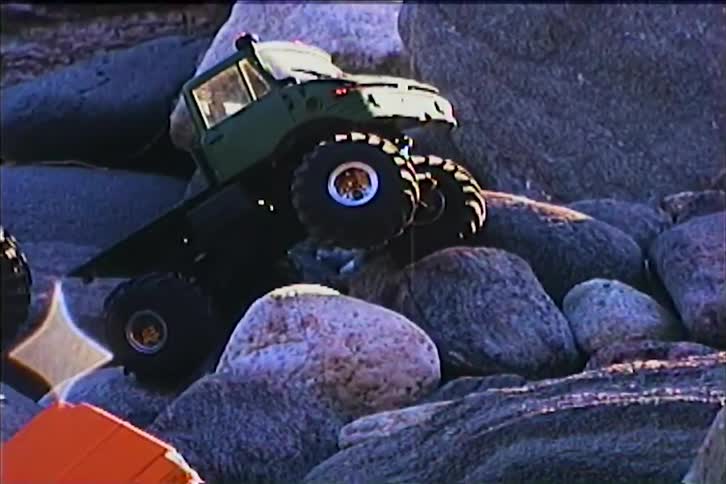 Several remote controlled monster trucks navigate a rocky and uneven terrain outdoors