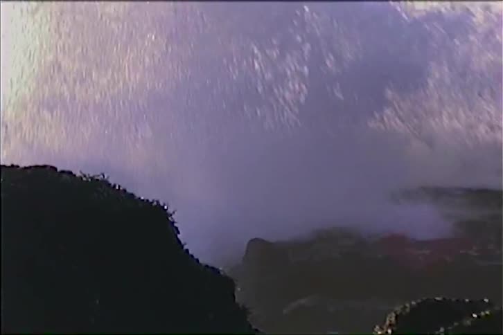 The first line of the video shows a static wide shot of an ocean landscape with dark jagged rocks silhouetted against the water and a pale cloudfilled sky the second line is a closeup of a powerful wave violently crashing against the rocks creating