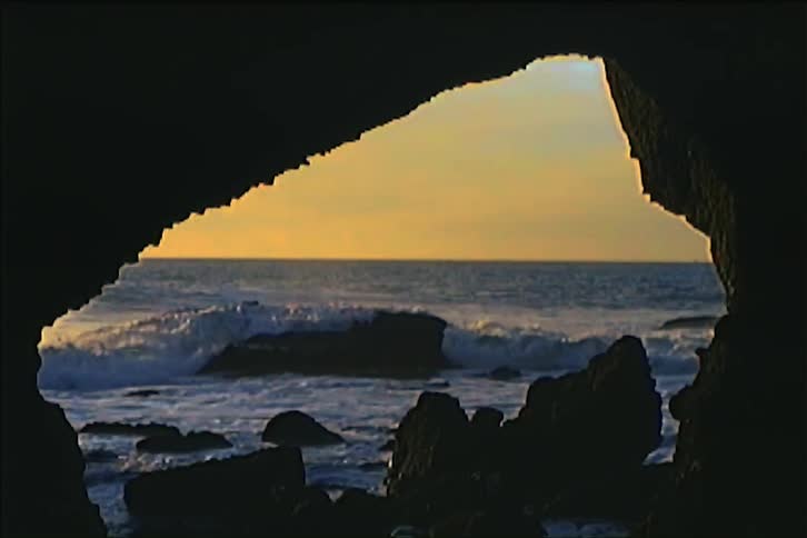 Ocean waves splash against rocks at the entrance of a coastal cave at sunset
