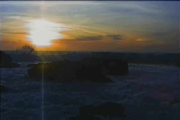 Ocean waves crashing aggressively against a rocky shoreline at sunset with the orange setting sun glowing in the sky