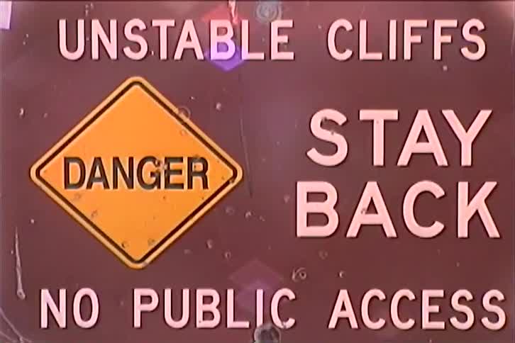 A sign on a cliff that reads unstable cliff stay back danger no public access with an ocean view beyond the cliff at sunset