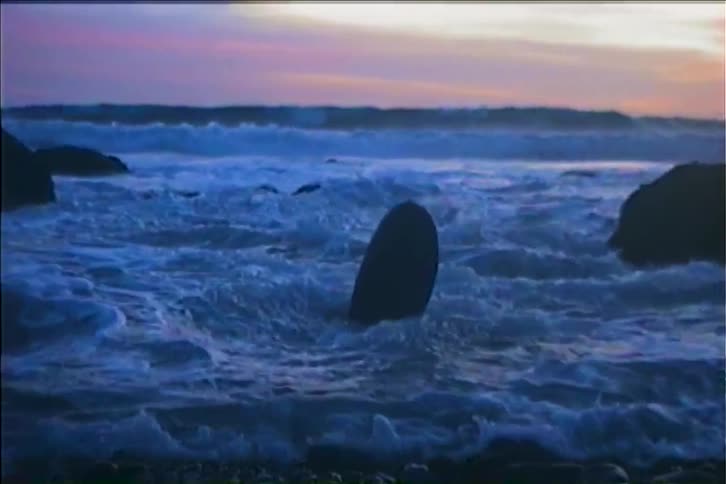 The first line shows a closeup of a surfboard standing upright on a rock on the coast the camera then slowly pulls back revealing a beautiful wide shot of the ocean with waves crashing on the shoreline as the surfboard remains perched on the rock