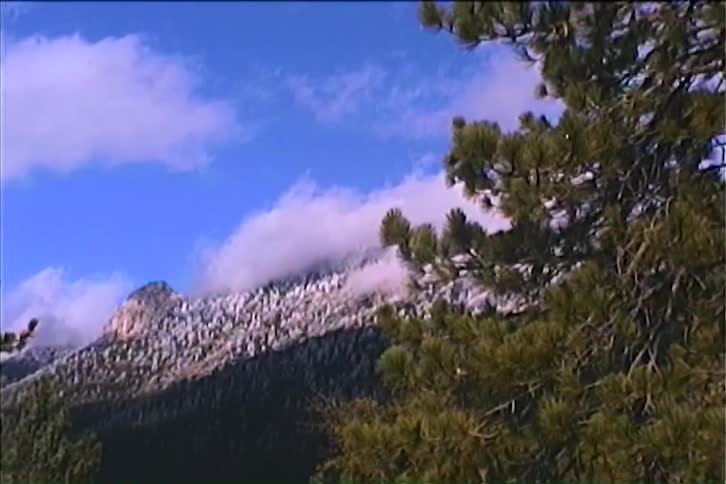 The video begins with a static wide shot of a snowcovered mountain landscape in the foreground there are the branches of pine trees covered with snow the camera then slowly zooms out and pans left revealing a wide beautiful view of the snowy mounta