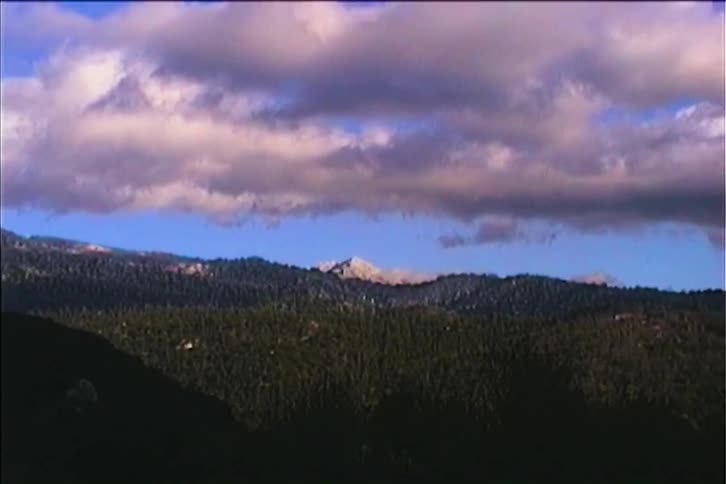 A long shot shows a snowcapped mountain peak nestled among a pine forest with puffy white and gray clouds floating in a brilliant blue sky above the camera slowly pans out revealing more of the vast mountain range and the forest below