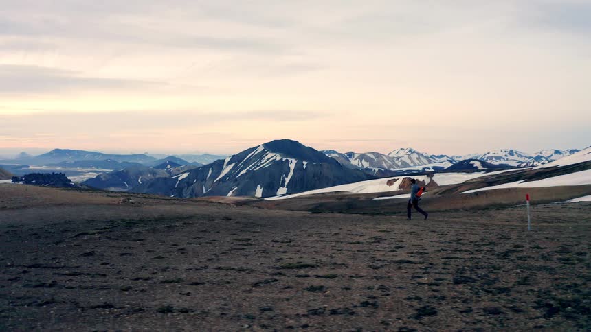 A lone hiker walks across a vast rocky plain towards snow capped mountains under a hazy sky