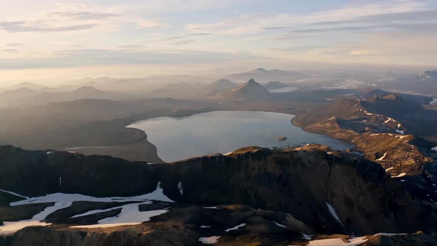 An aerial shot shows a wide mountainous landscape with some patches of snow and ice a large beautiful lake sits between the peaks as the sun casts a hazy golden light across the landscape