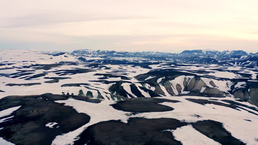 This drone video captures a wide shot of a vast snowcovered landscape the camera slowly pans to the right revealing more of the rugged terrain and distant mountains under a dusky sky