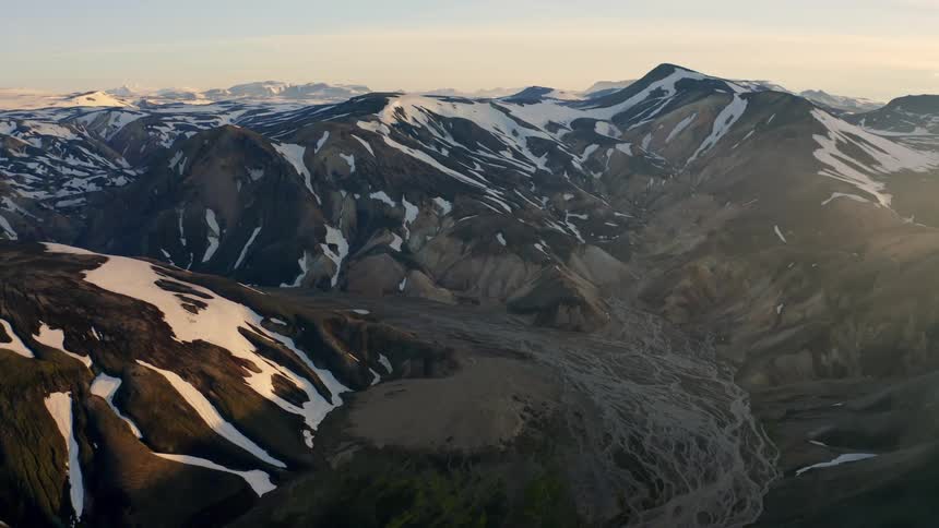 The first line shows a majestic wide aerial shot of a vast mountain range with snowy peaks and deep valleys the second line shows the camera slowly moving forward over the landscape revealing the rugged textured terrain and a dry riverbed