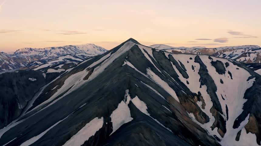 This aerial video captures a striking mountain peak in a vast snowcovered landscape the camera pulls back revealing a broader view of the mountain range as the sky transitions from sunset hues to a softer light