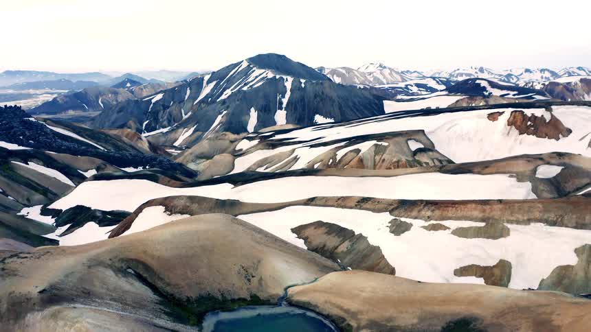 An aerial shot captures a vast mountain landscape with peaks covered in snow and ice the camera glides past the mountains revealing a small turquoisecolored glacial lake at their base