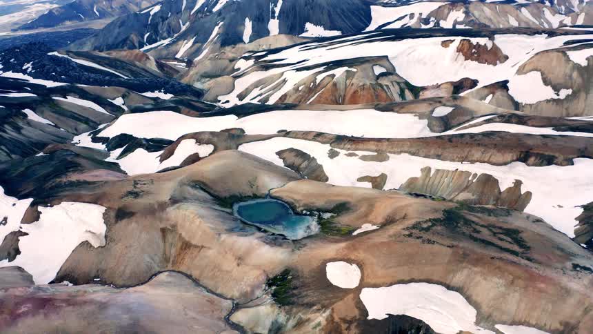 The first line shows a beautiful wide aerial shot of a majestic mountainous landscape with jagged peaks partially covered in snow the second line shows the camera slowly panning down and to the right revealing a small light blue glacial lake nestled