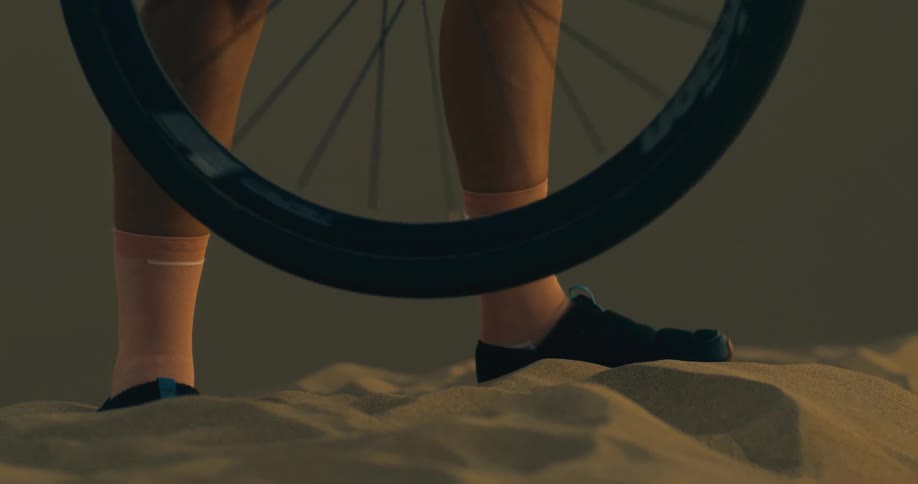 A persons feet standing on sand with a bicycle wheel spinning above the sand until it slows stops and gently starts spinning in the opposite direction