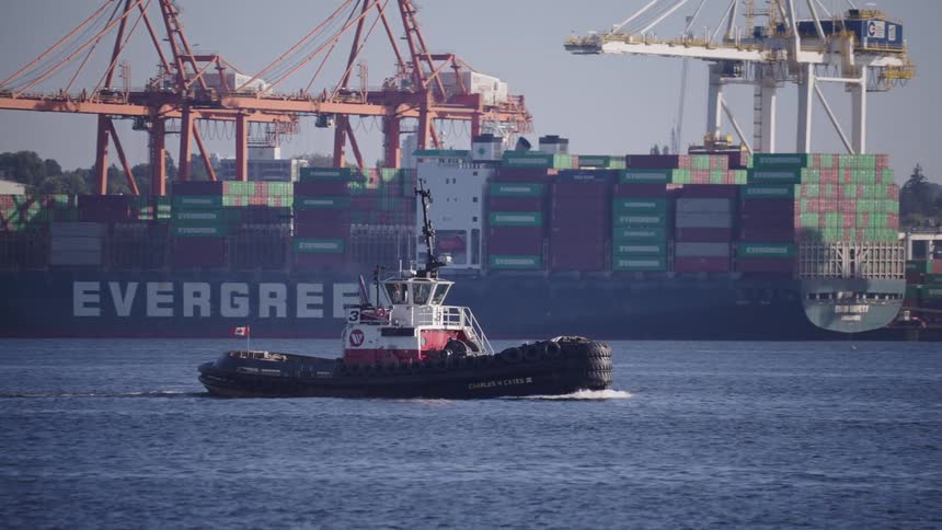 Tugboat sails in front of a port with cranes and containers