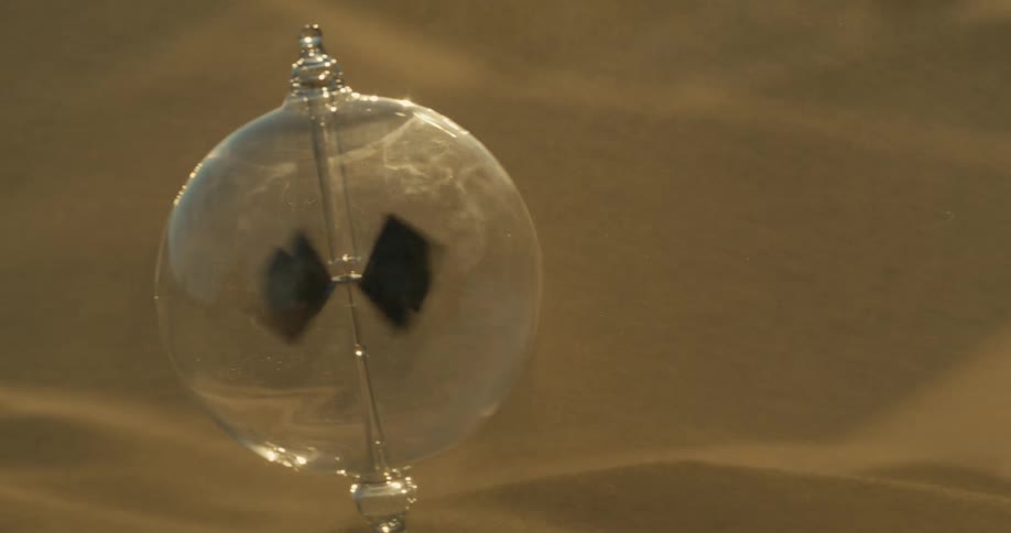 A radiometer with four vanes inside a glass bulb rotates on a sand surface illuminated by a light source