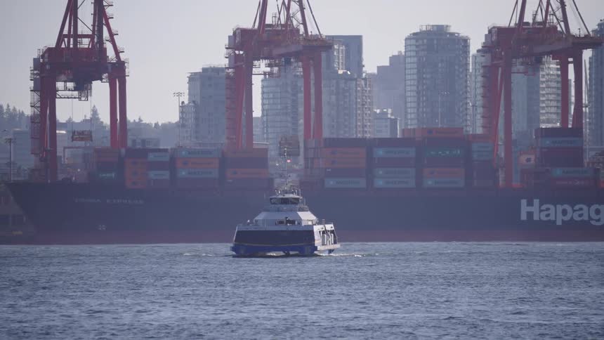 Cargo ship with containers docked at a port with cranes and a ferry sailing in the foreground