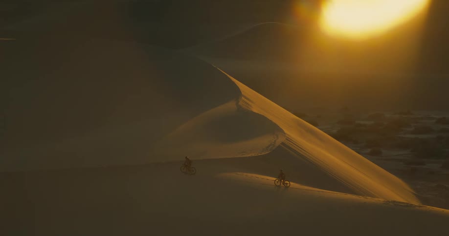 Aerial of two people moving their bicycles across a sand dune in the desert at sunset