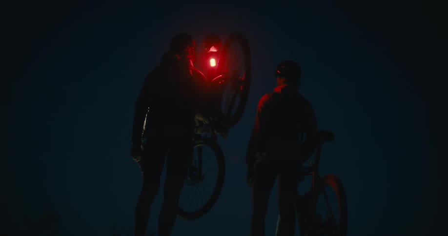 Male cyclist picks up his bicycle while his friend continues to push their bike as they both walk at night