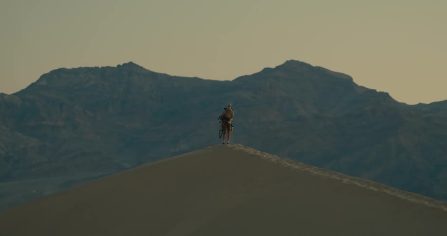 A person on a sand dune is holding a bike and looks at the mountains in the background
