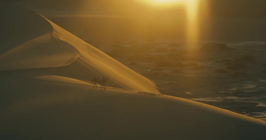 Two people walking across sand dunes in a desert during daytime while carrying their bicycles
