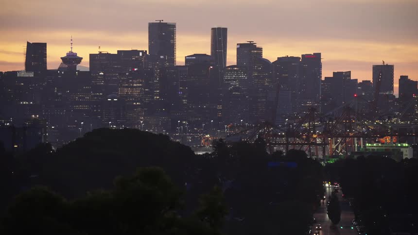 A long shot captures a city skyline at dusk with lights twinkling on the highrise buildings below the city a highway is filled with cars their headlights creating streaks of light as they move along the road