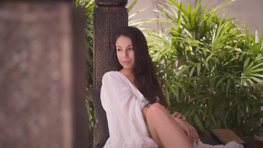 A young woman wearing a robe sits on wooden structure with plants in the background on a sunny day
