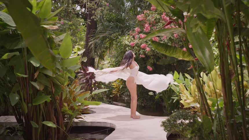 Woman in a white swimsuit spinning with a coverup among blooming tropical vegetation