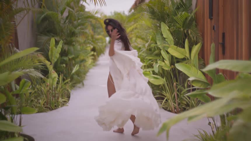 Woman wearing a flowing outfit and sunglasses passes large wooden doors while running along a path bordered by tropical plants on a summer day