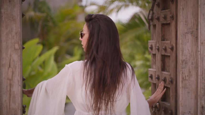 This video shows a woman with her back to the camera standing in front of a pair of large intricately carved wooden doors she reaches out and opens the doors revealing a lush green tropical garden before turning and smiling at the camera
