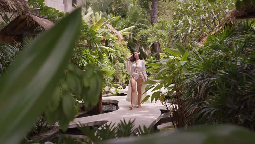Woman walks in a white bikini and light coverup along a path surrounded by tropical greenery