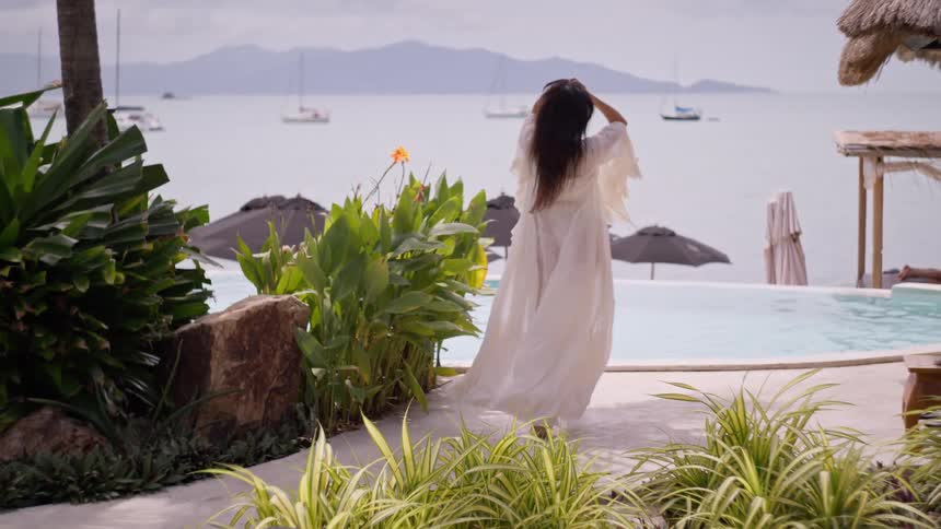 Woman in a white dress walks smiling near the pool with an ocean view