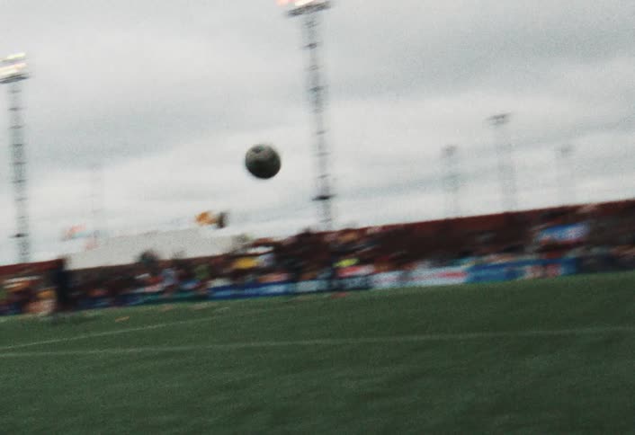 A person standing on a field kicks a ball toward a goal and the ball goes high into the air as people watch from the stands