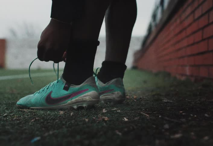 Player on the field tying a blue soccer cleat with pink details next to a red brick wall