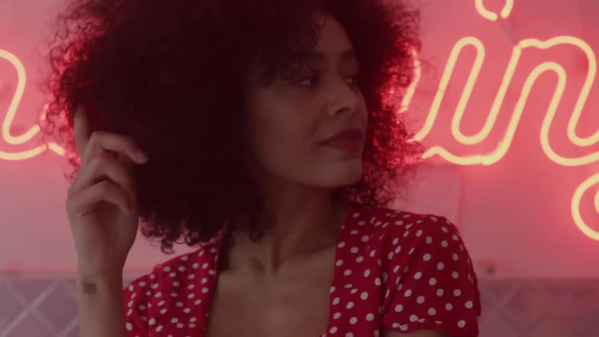 A woman with voluminous curly hair in a red polka dot dress poses playfully against a pink neon sign smiling at the camera