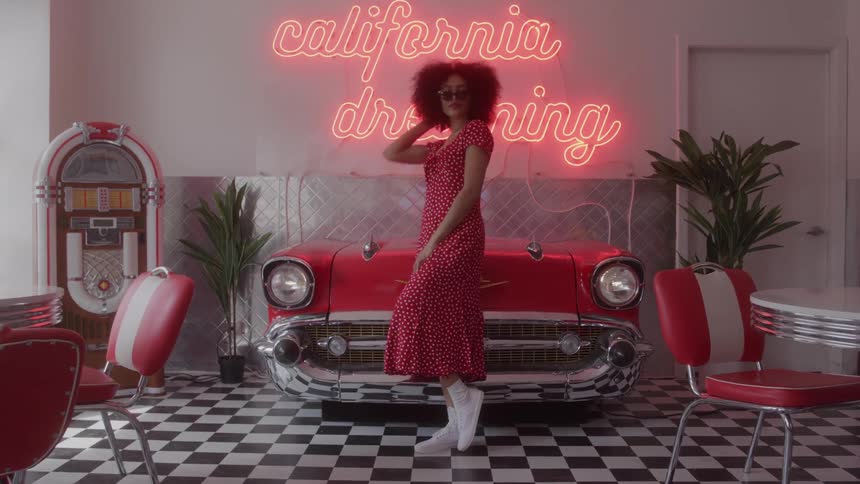 A woman in a red polka dot dress and sunglasses poses in a retro diner setting with a red vintage car and neon sign