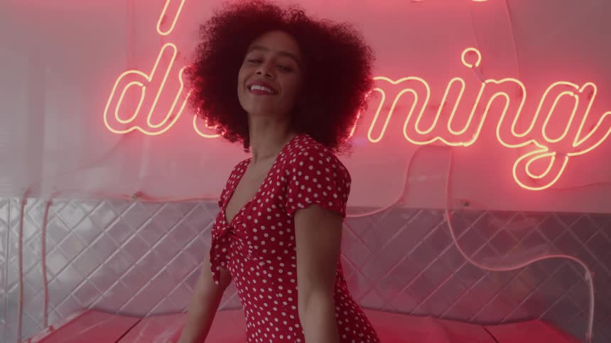 A woman stands in front of a neon sign in an indoor setting smiling and moving her body while looking toward the camera