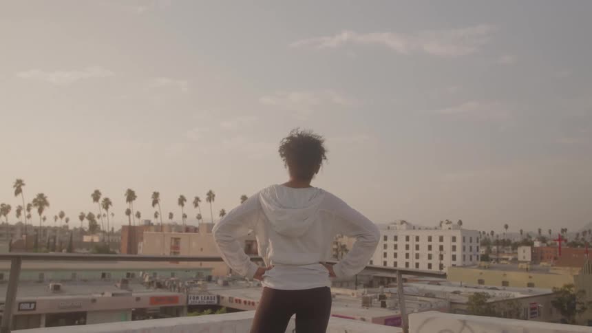 A person with their back to the camera stands on a rooftop looking out over a city theyre wearing a white hoodie and have their hands on their hips